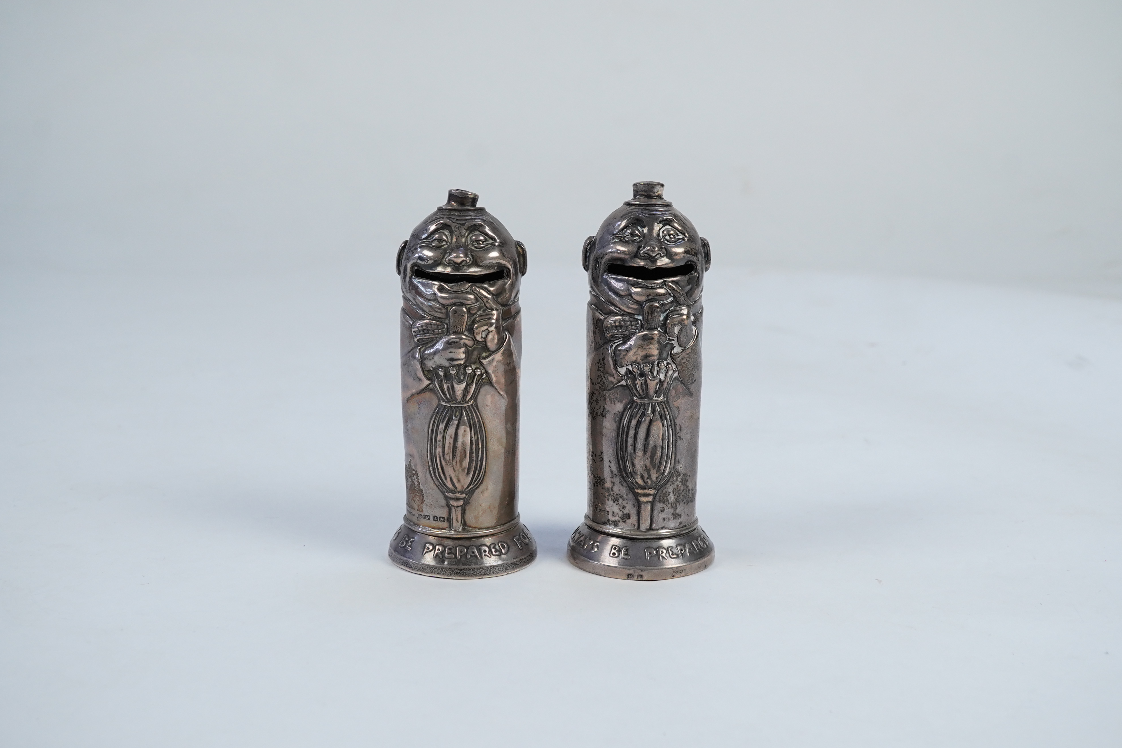 A matched pair of George V novelty silver money boxes, each cylindrical box embossed as a banker with an umbrella, the circular base inscribed 'Always be prepared for a rainy day'